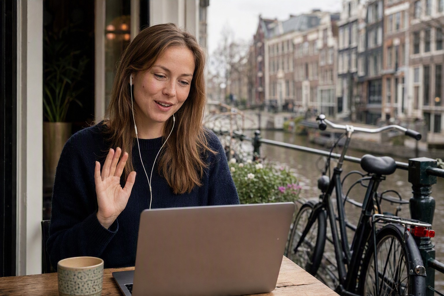 A woman wearing earphones waves at her laptop during a voice call at a canal-side table, with Dutch canal houses and parked bicycles behind her