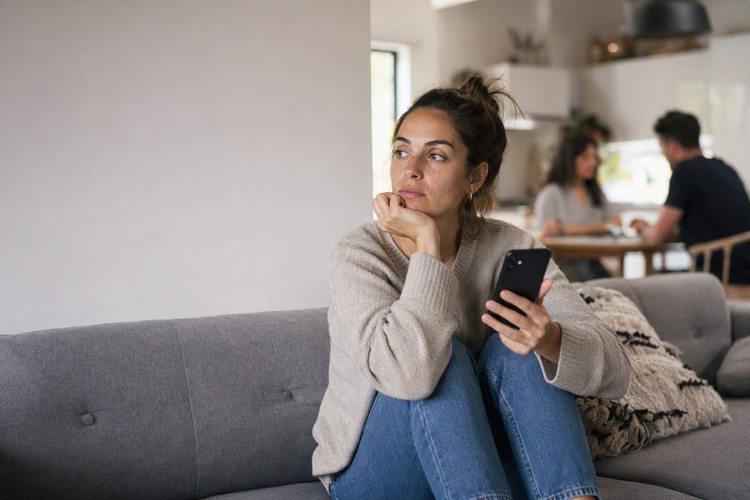 A woman on a couch holding her phone, looking away in thought while others sit together in the background